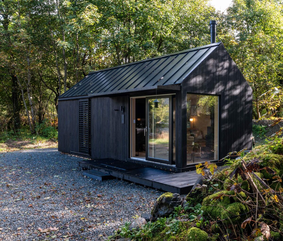 A dark timber cabin sits among trees, its metal roof visible above a gravel drive lined with rocks.